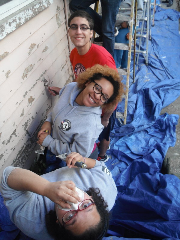 Two AmeriCorps members scraping paint from the side of a house